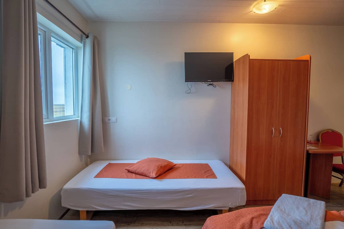 A single bed with a white mattress and orange bedding, a wooden wardrobe, a television mounted on the wall, and a small table with a chair.