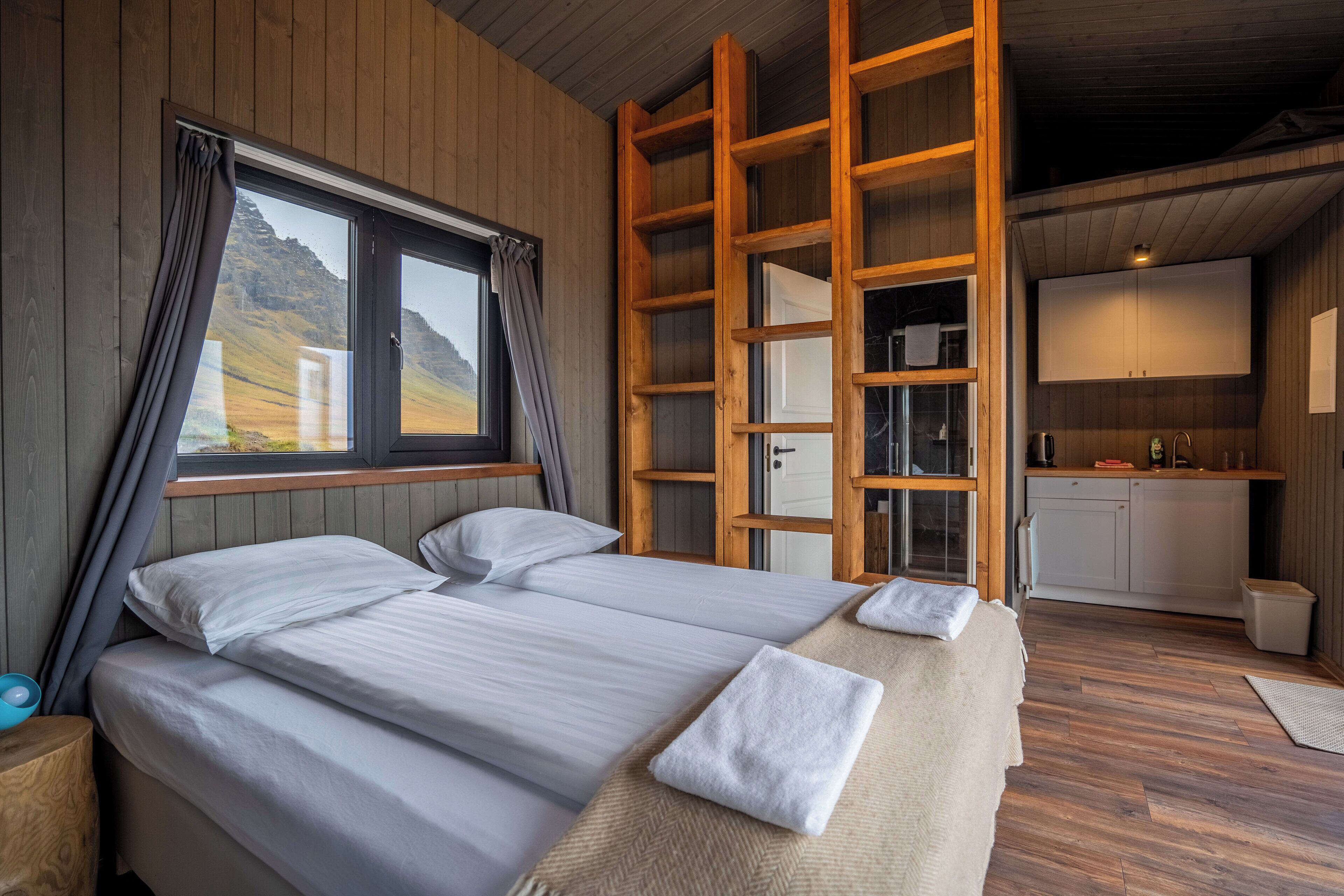 A bedroom with two beds, a wooden ladder, a window with curtains, a view of the outdoors, and a small kitchenette.