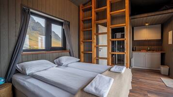 A bedroom with two beds, a wooden ladder, a window with curtains, a view of the outdoors, and a small kitchenette.