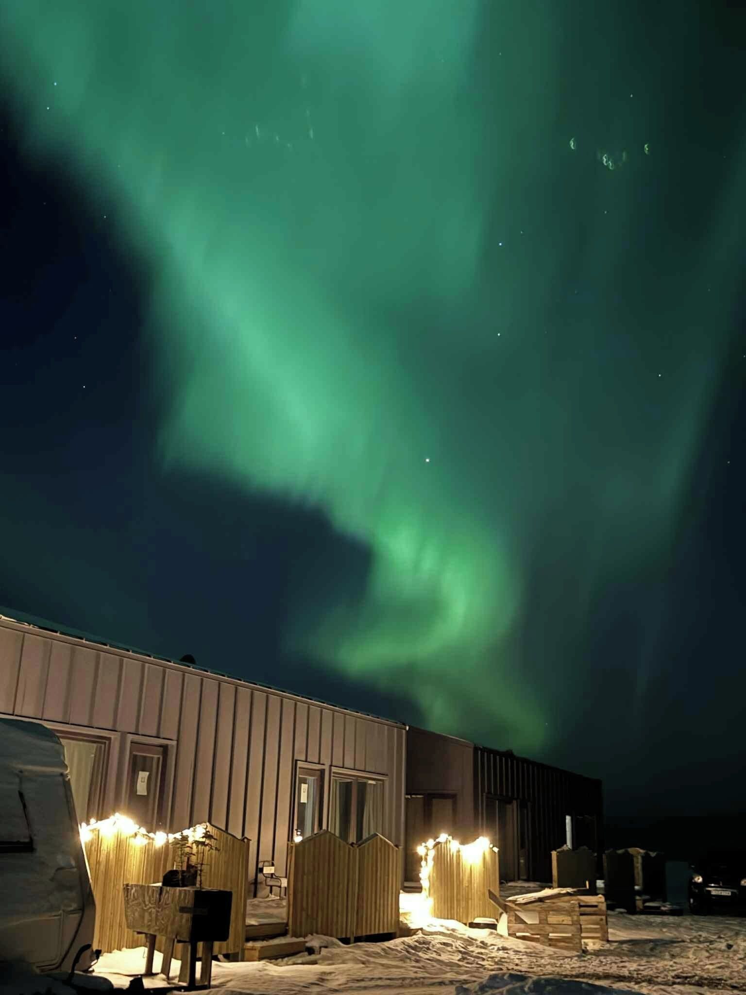 En bygning med snedækket jord, trækonstruktioner og udsigt til nattehimmelen med grønne nordlys.