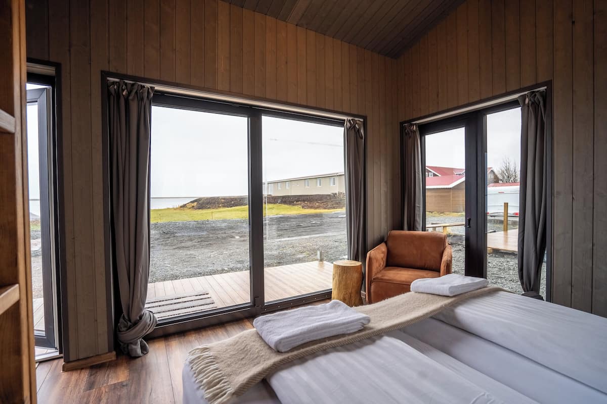 A room with a large bed, a wooden floor, and a view of a building and a beach through large windows.