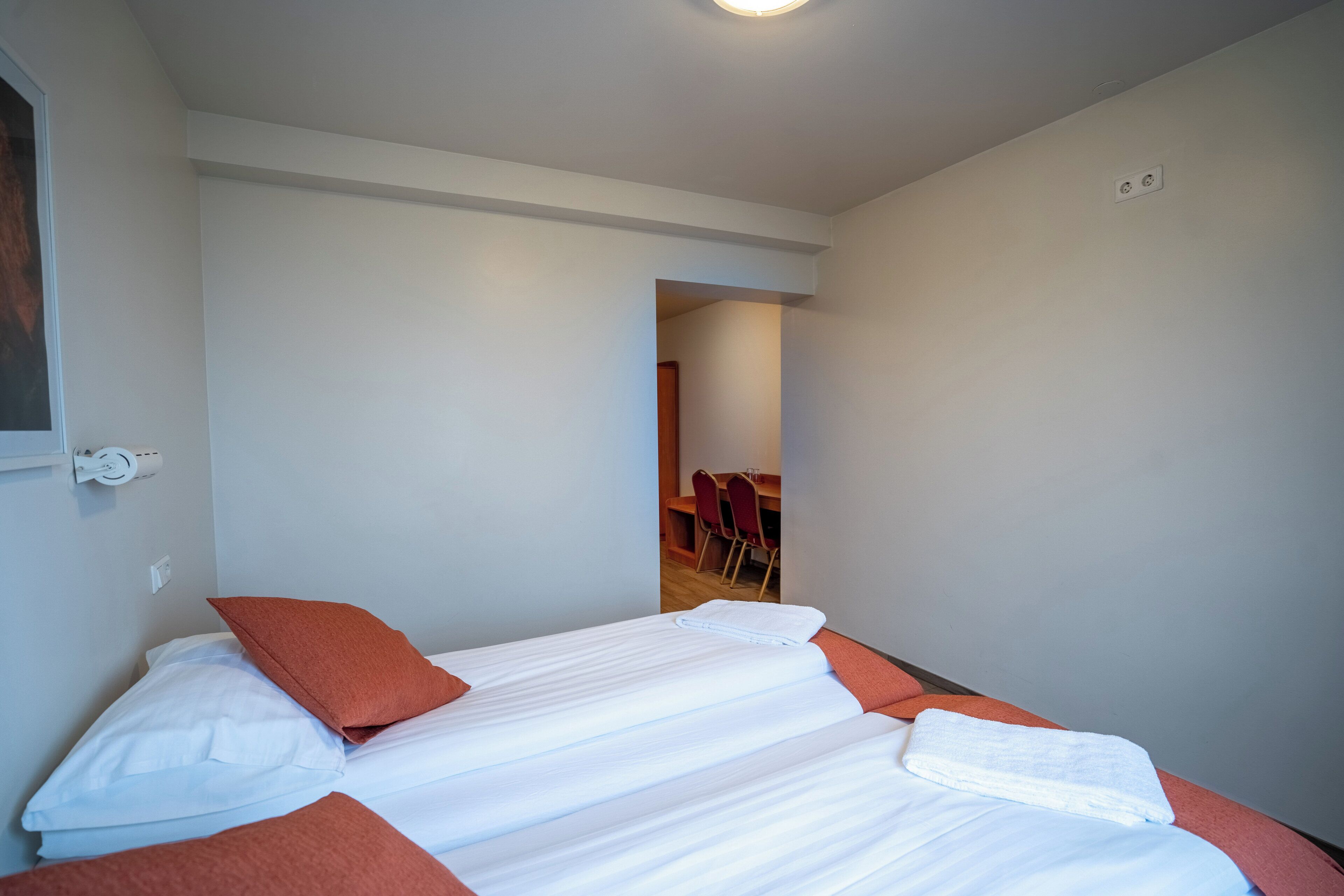 A single bed with white linens and orange pillows, a small table with a chair, and a door leading to a dining area.