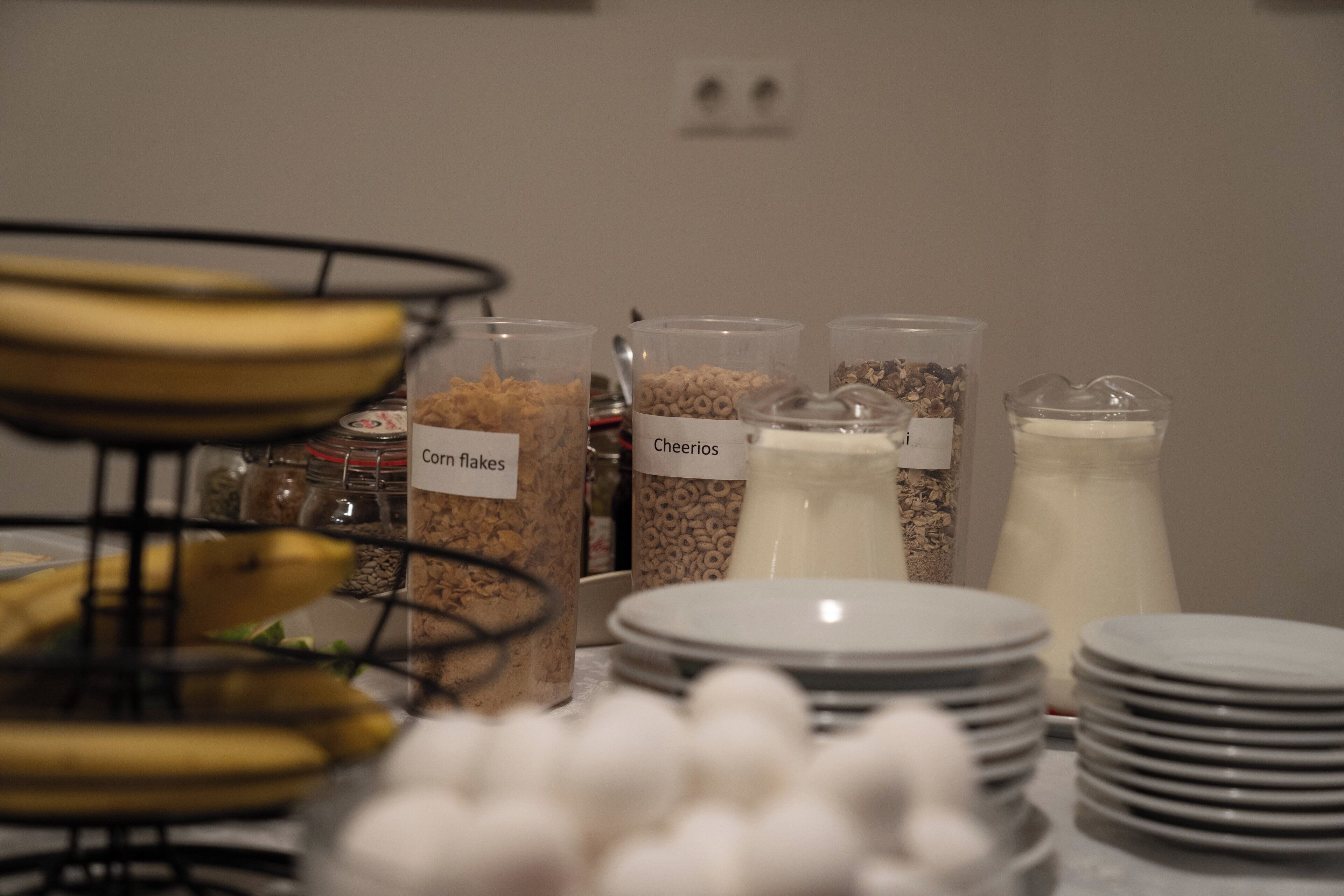 A breakfast buffet with labeled containers of cereal and milk, and a banana stand.