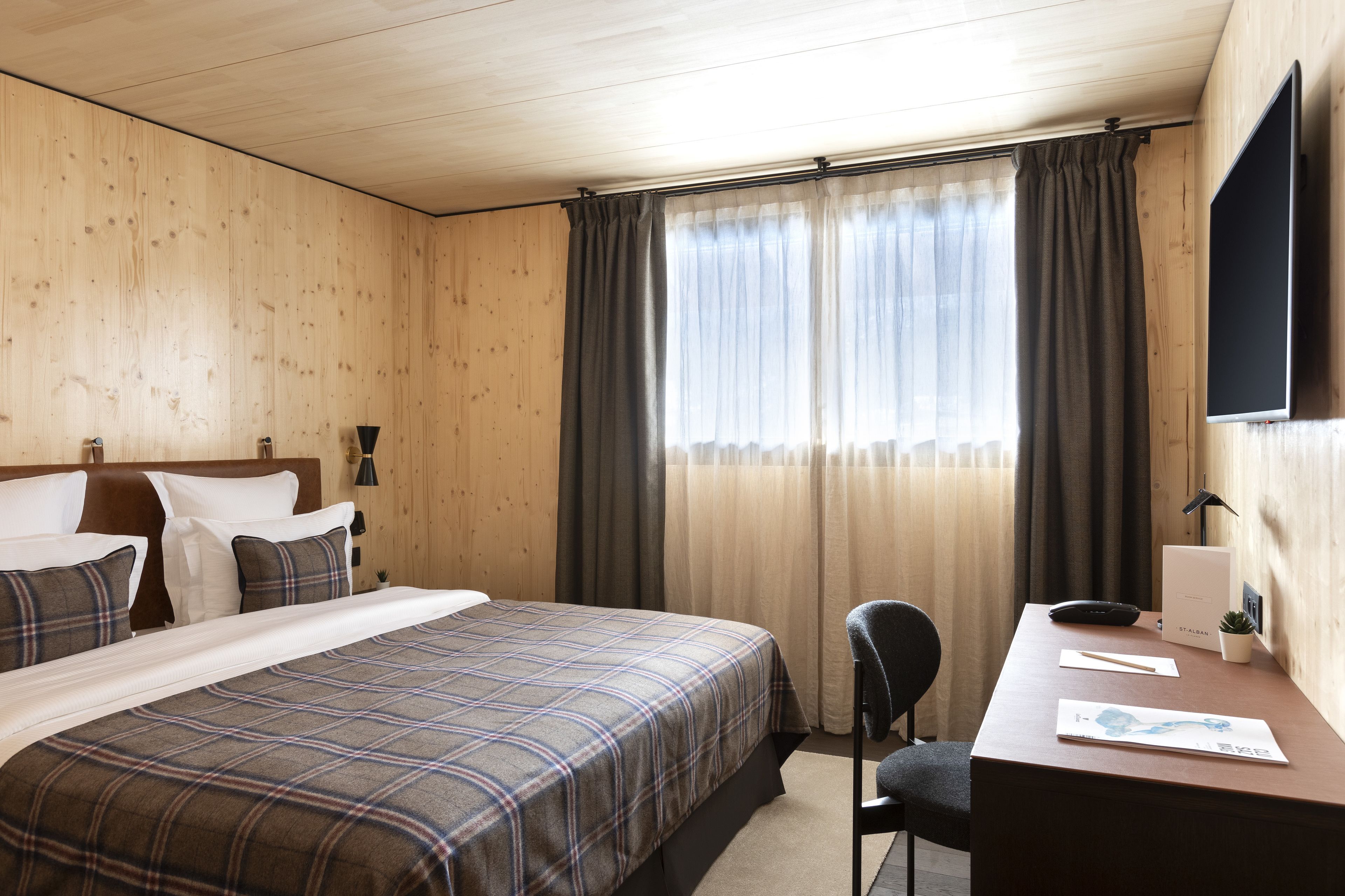 Een hotelkamer met een houten plafond, een bed met een geruit deken, een bureau met een stoel, een flatscreen-televisie en een raam met gordijnen.