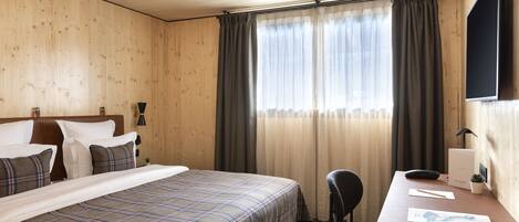 Een hotelkamer met een houten plafond, een bed met een geruit deken, een bureau met een stoel, een flatscreen-televisie en een raam met gordijnen.