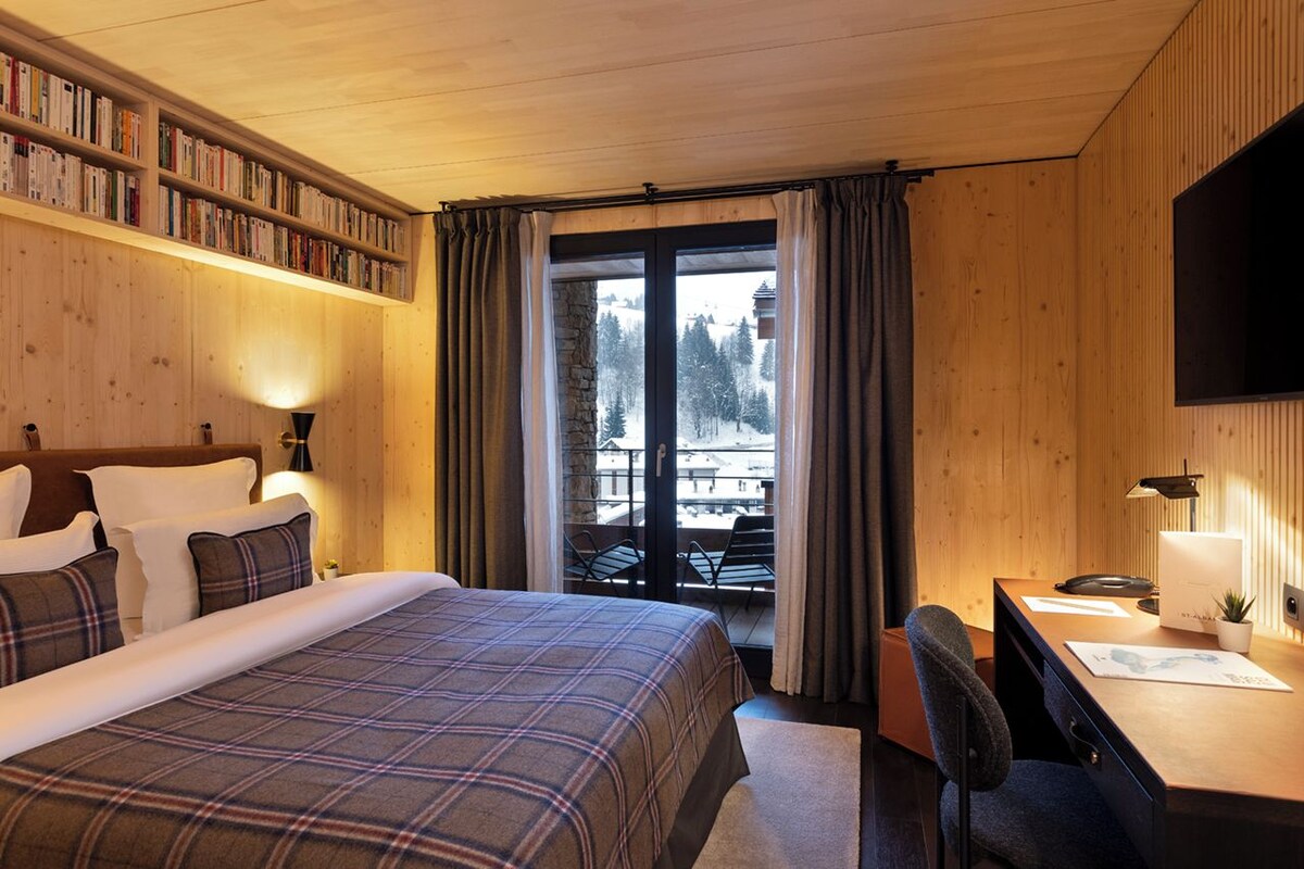 Habitación de hotel con una cama grande, un escritorio, una estantería y un balcón con vistas a árboles.