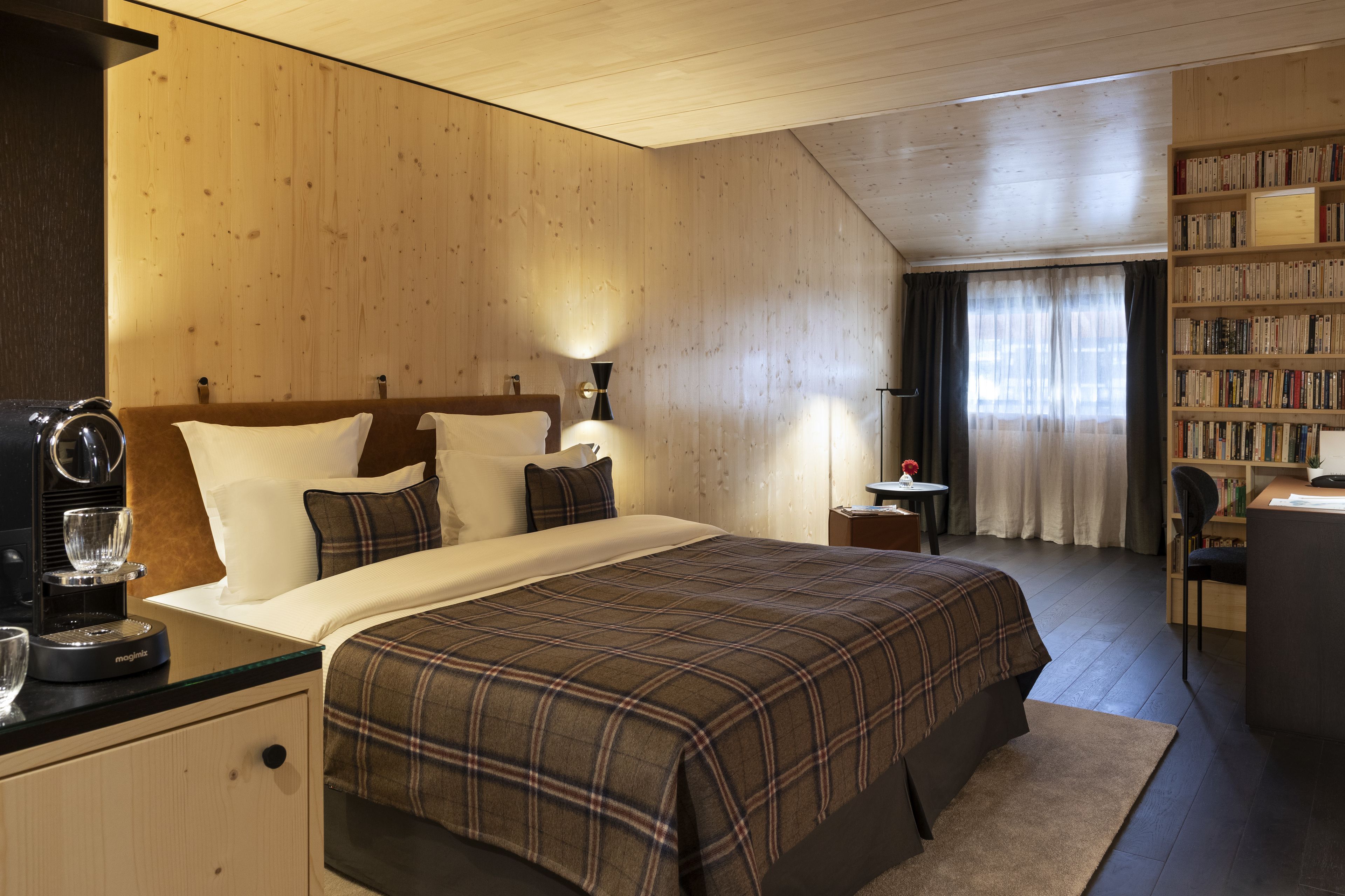 A hotel room with a wooden ceiling, a large bed with a plaid bedspread, a bedside table with a coffee maker, a bookshelf, and a window with curtains.