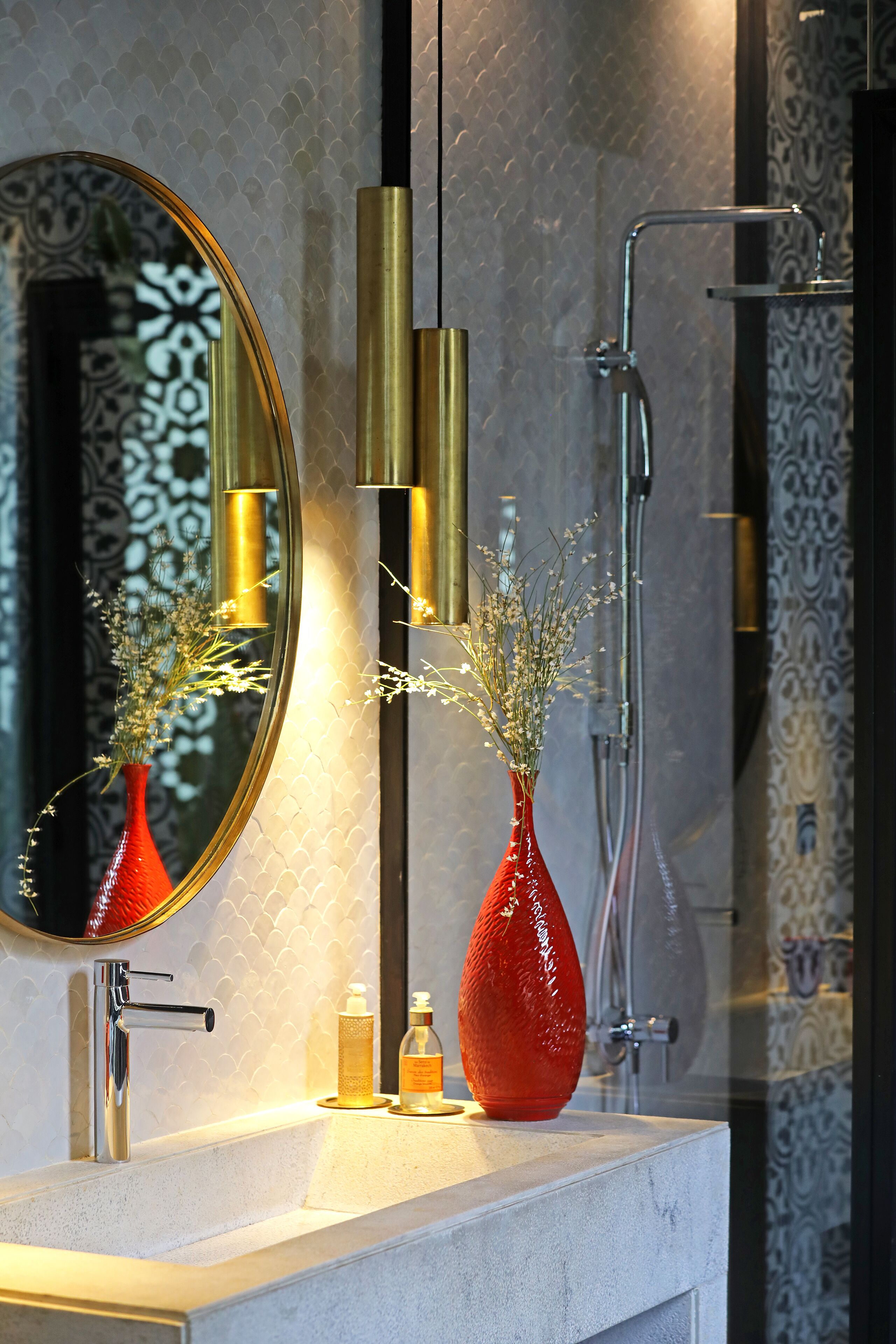 Un baño con espejo, un jarrón rojo con flores, grifo y ducha.