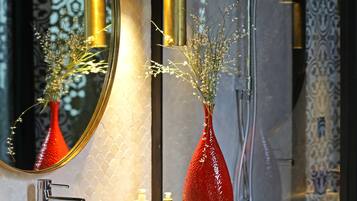 Un baño con espejo, un jarrón rojo con flores, grifo y ducha.