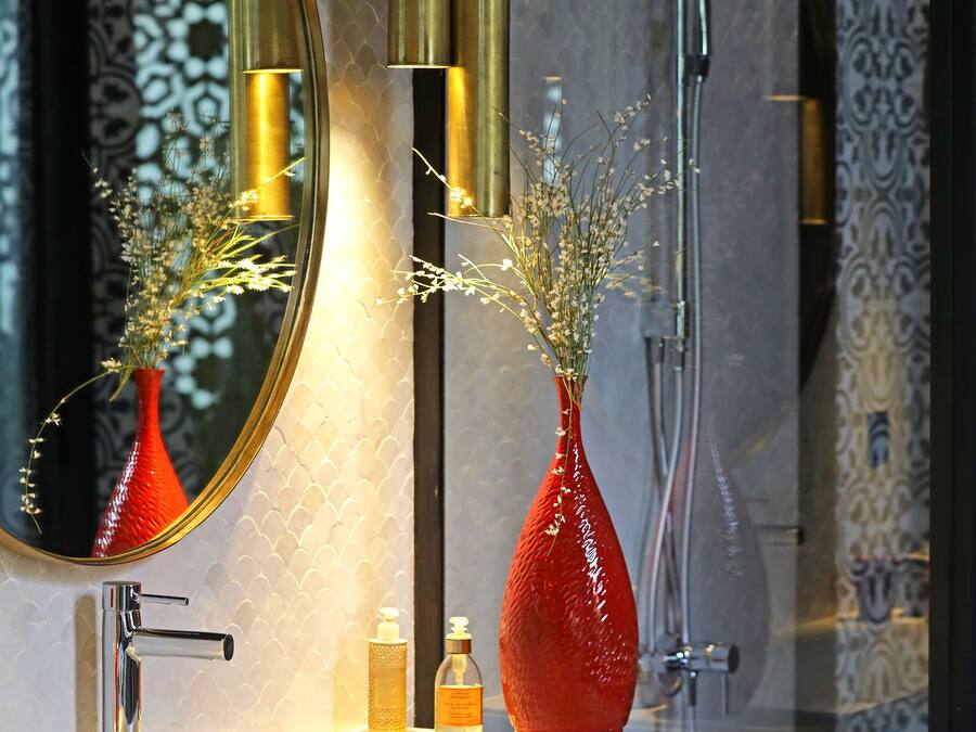 Un baño con espejo, un jarrón rojo con flores, grifo y ducha.