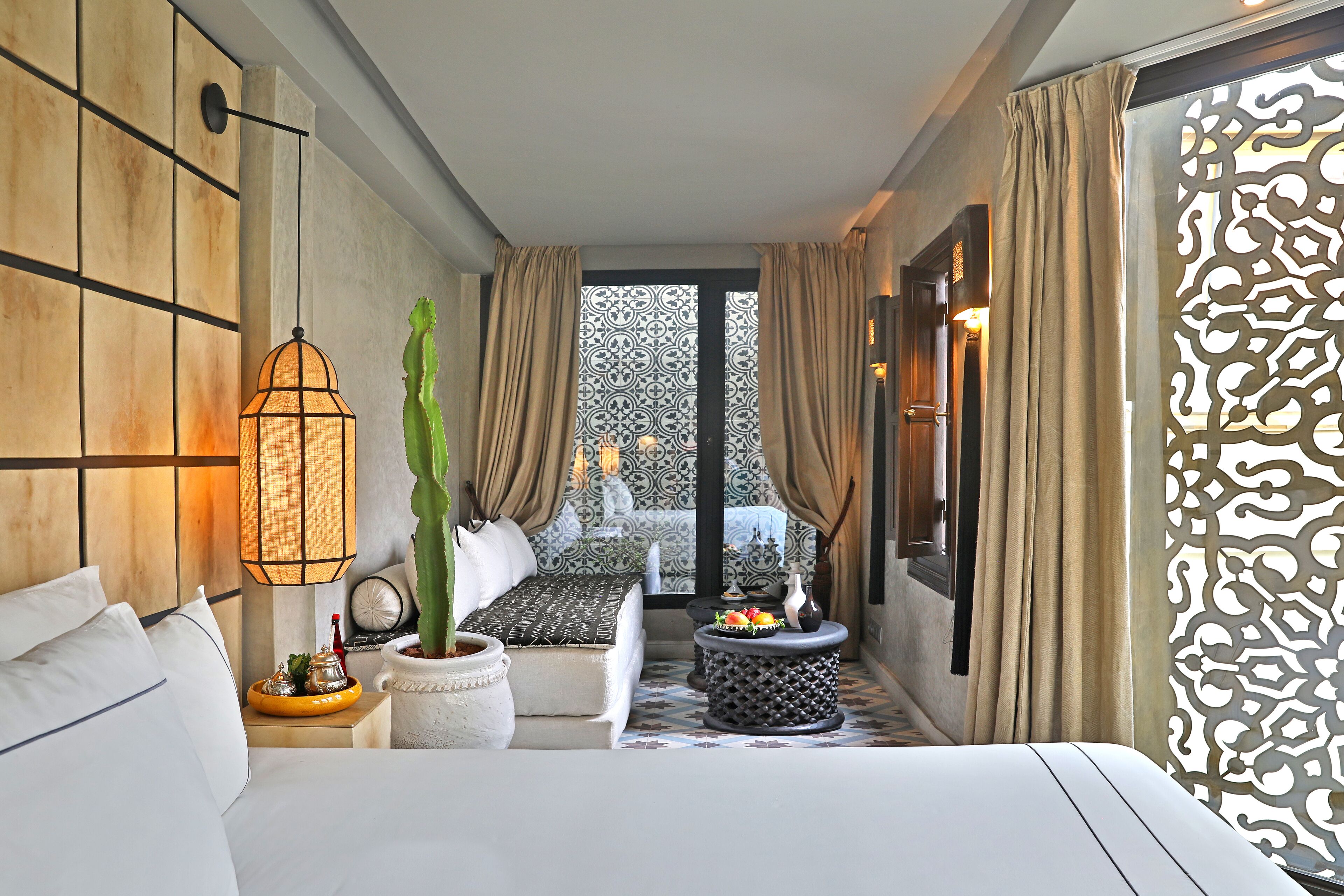 Une chambre d’hôtel avec un lit, une table de chevet avec une lampe, une plante en pot, une petite table ronde agrémentée d’un objet décoratif, et une vue à travers une porte à motifs.