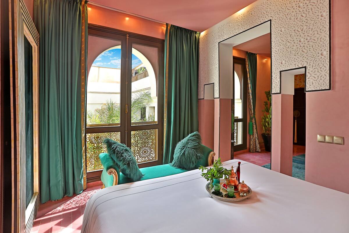 Ein Hotelzimmer mit einem großen Bett, einem Fenster mit grünen Vorhängen, einem Zier-Spiegel und einer Ablage mit Gegenständen auf dem Bett.