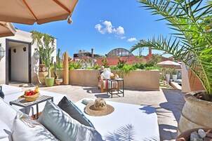 A rooftop patio with a white sofa, a small table with a basket of fruit, and a straw hat on the seat.