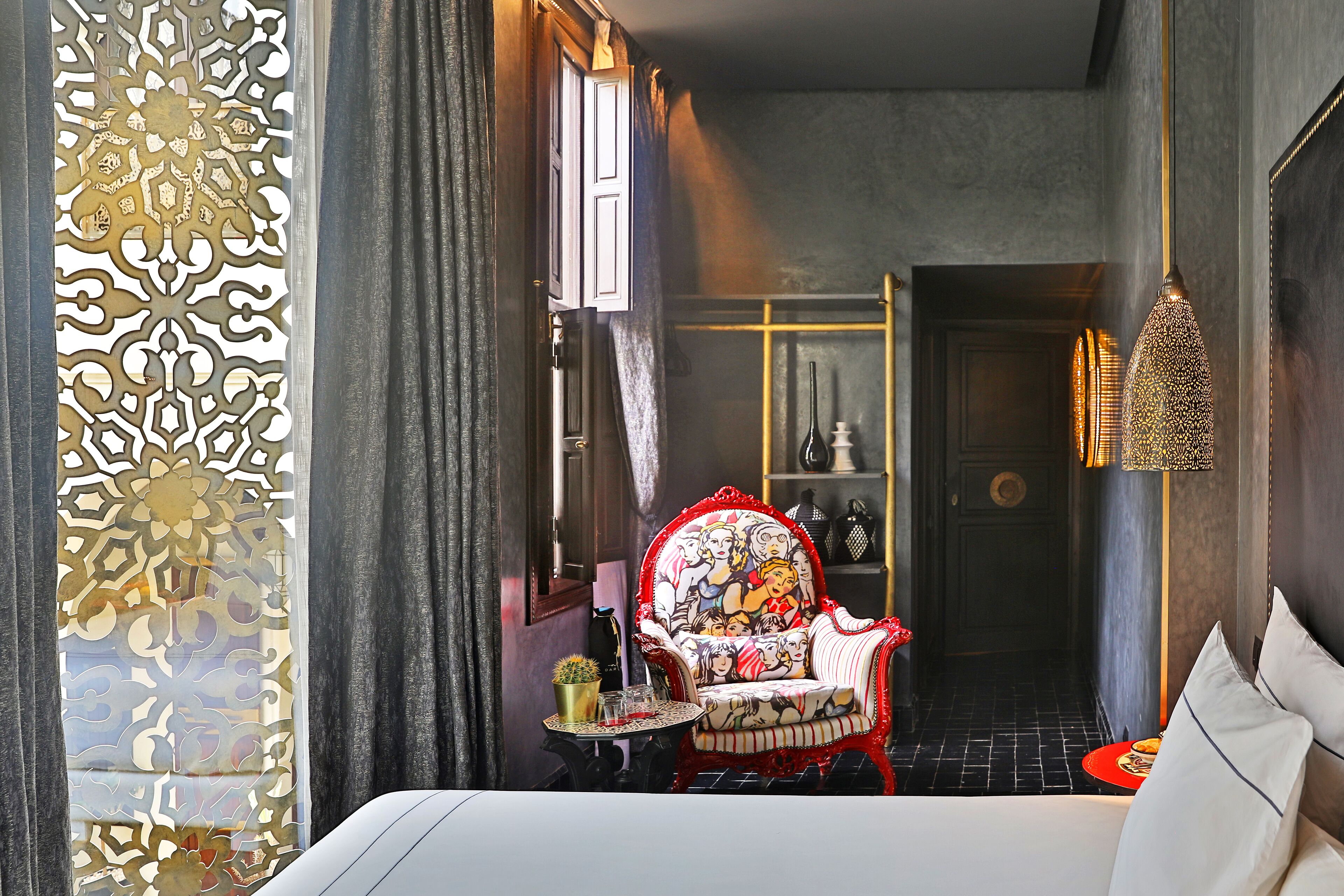 A hotel room with a bed, a decorative chair, a small table with a plant, and a door leading to another room.