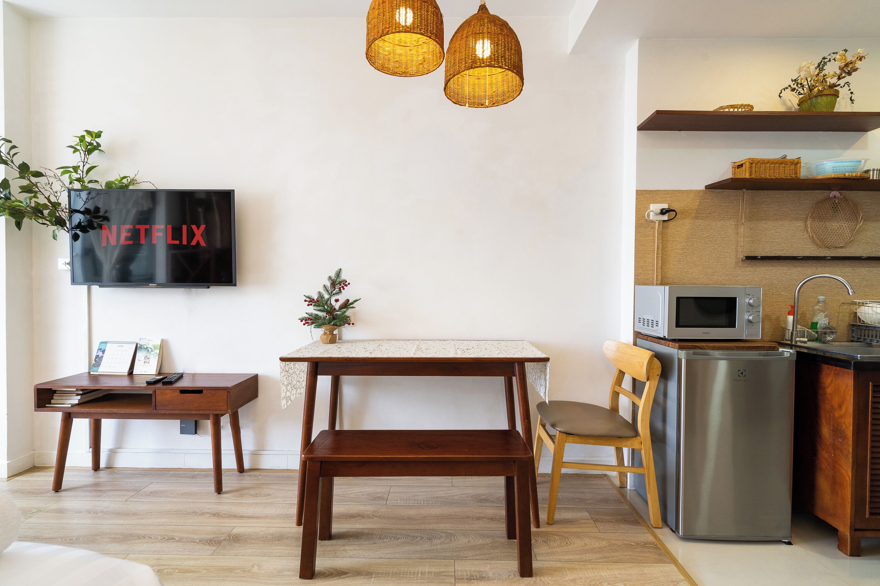 Un salon moderne avec un bureau en bois, un téléviseur, des luminaires suspendus en osier et un coin cuisine équipé d’un four à micro-ondes et d’un réfrigérateur.