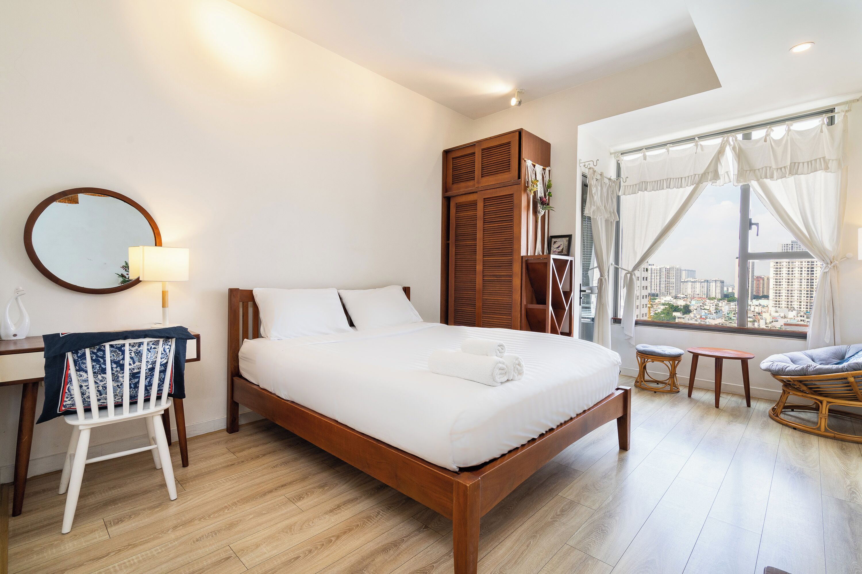 A modern hotel room with a wooden bed, a round mirror, a desk, and a view of the city through large windows.