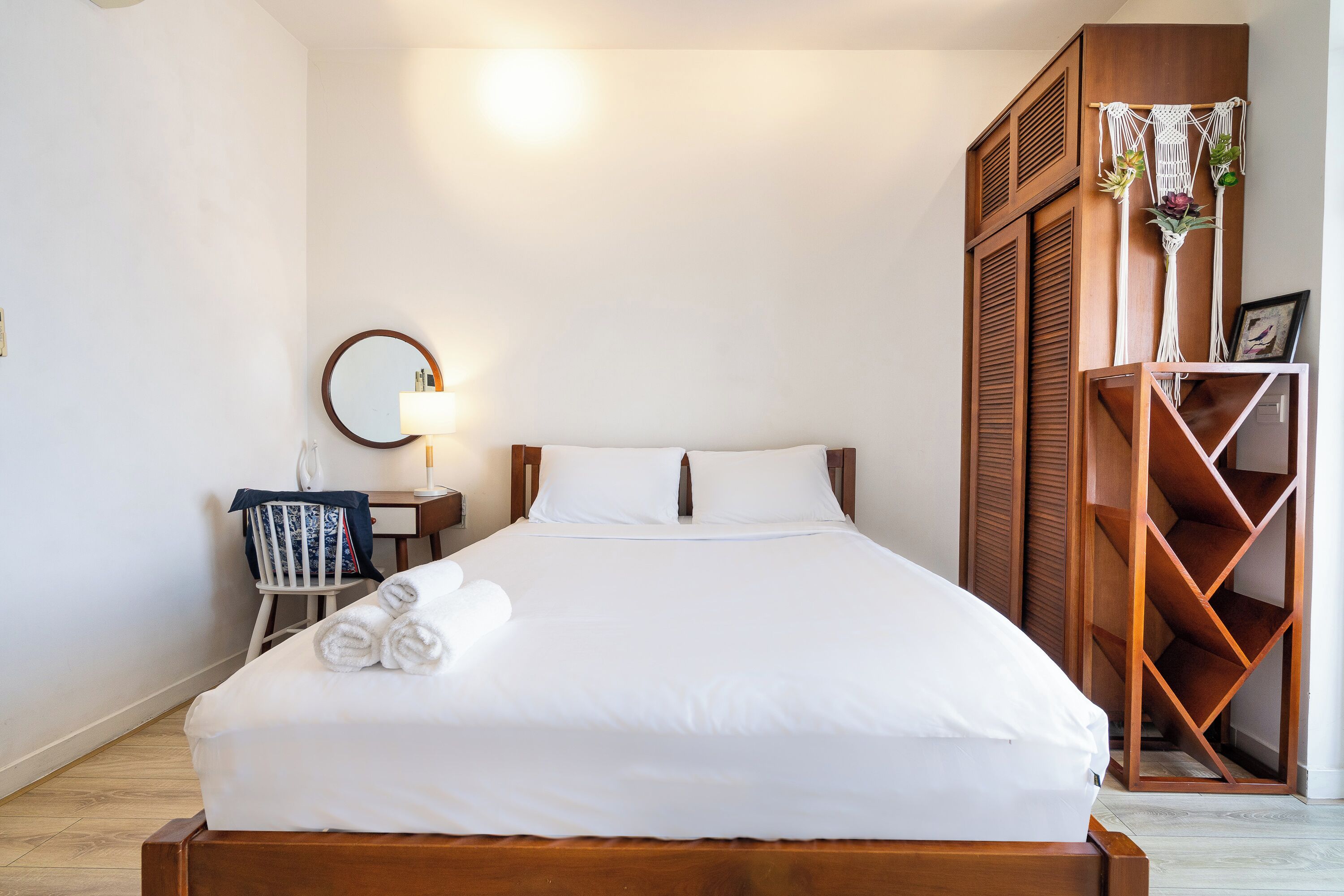 A neatly made bed with white linens, a bedside table with a lamp, and a wooden wardrobe with a decorative plant.