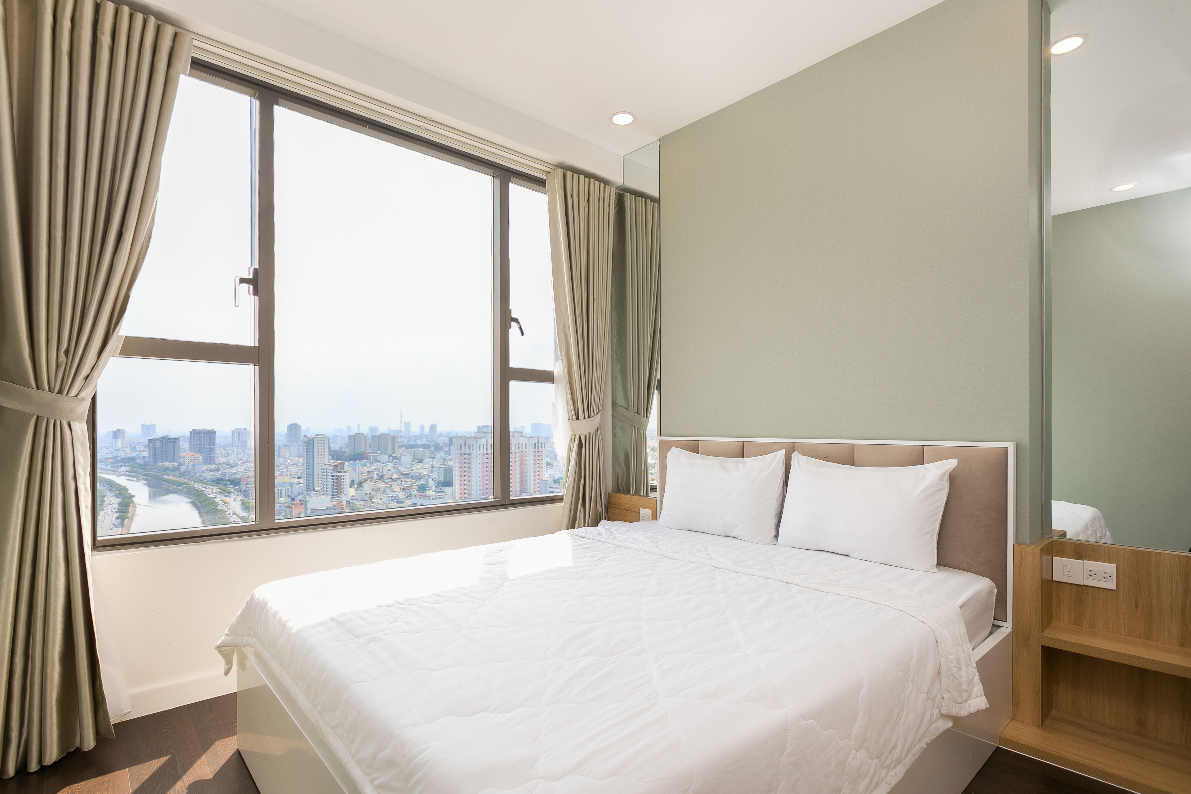 Una habitación de hotel con una cama grande, vistas de la ciudad a través de una ventana y una mesita de noche de madera.