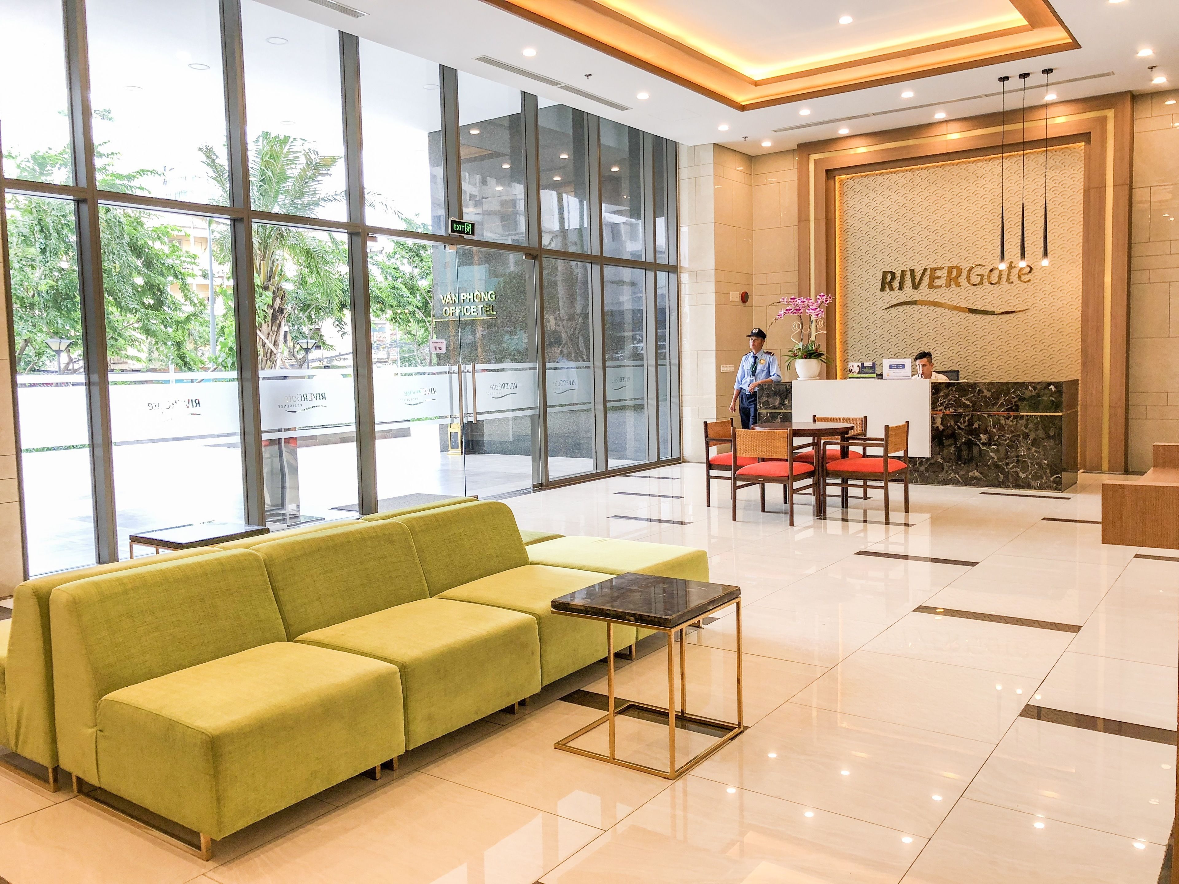 A hotel lobby with a reception desk, a green sofa, and a large window offering a view of the outside.