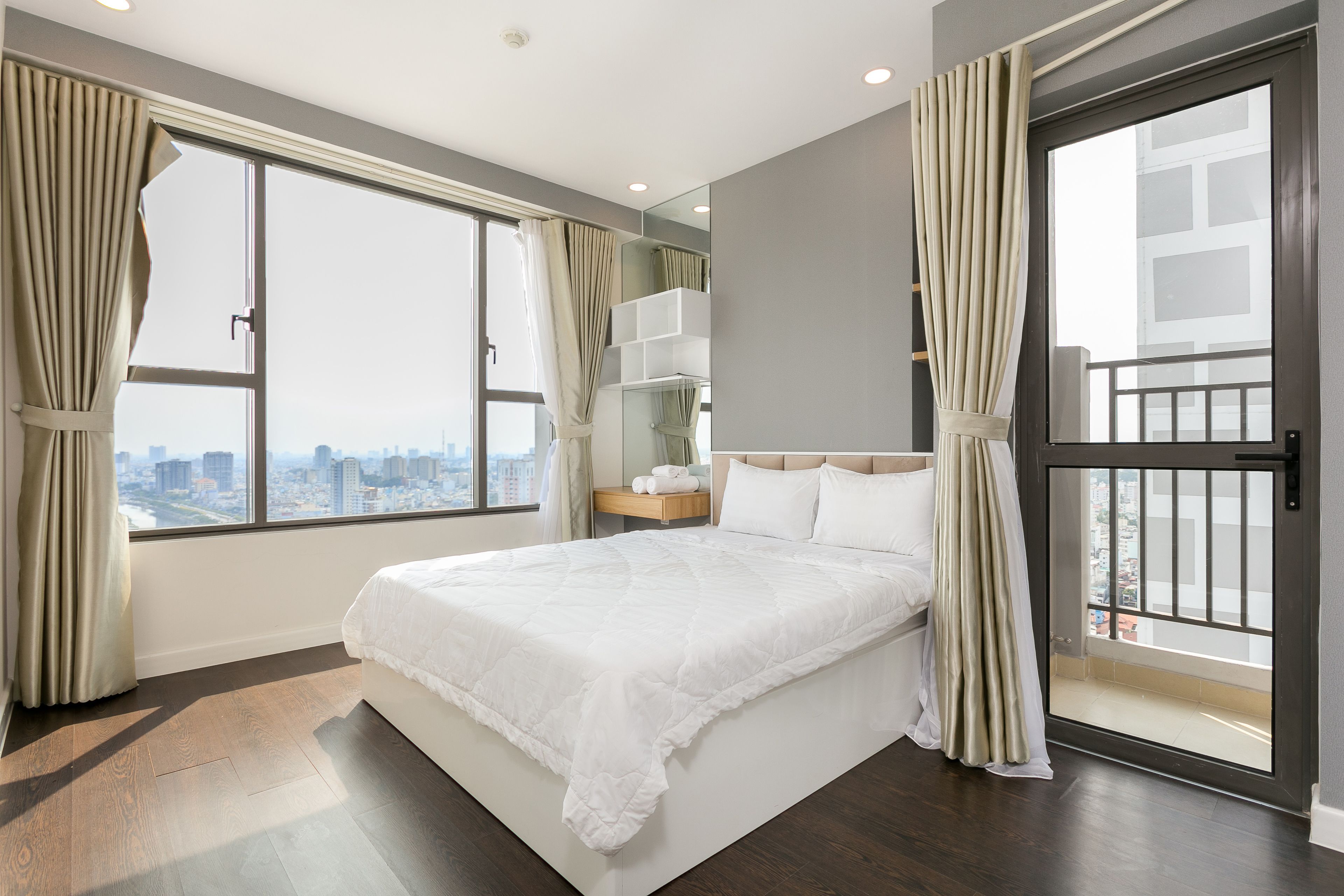 A modern bedroom with a large bed, a nightstand, a mirror, and a view of the city through a large window.