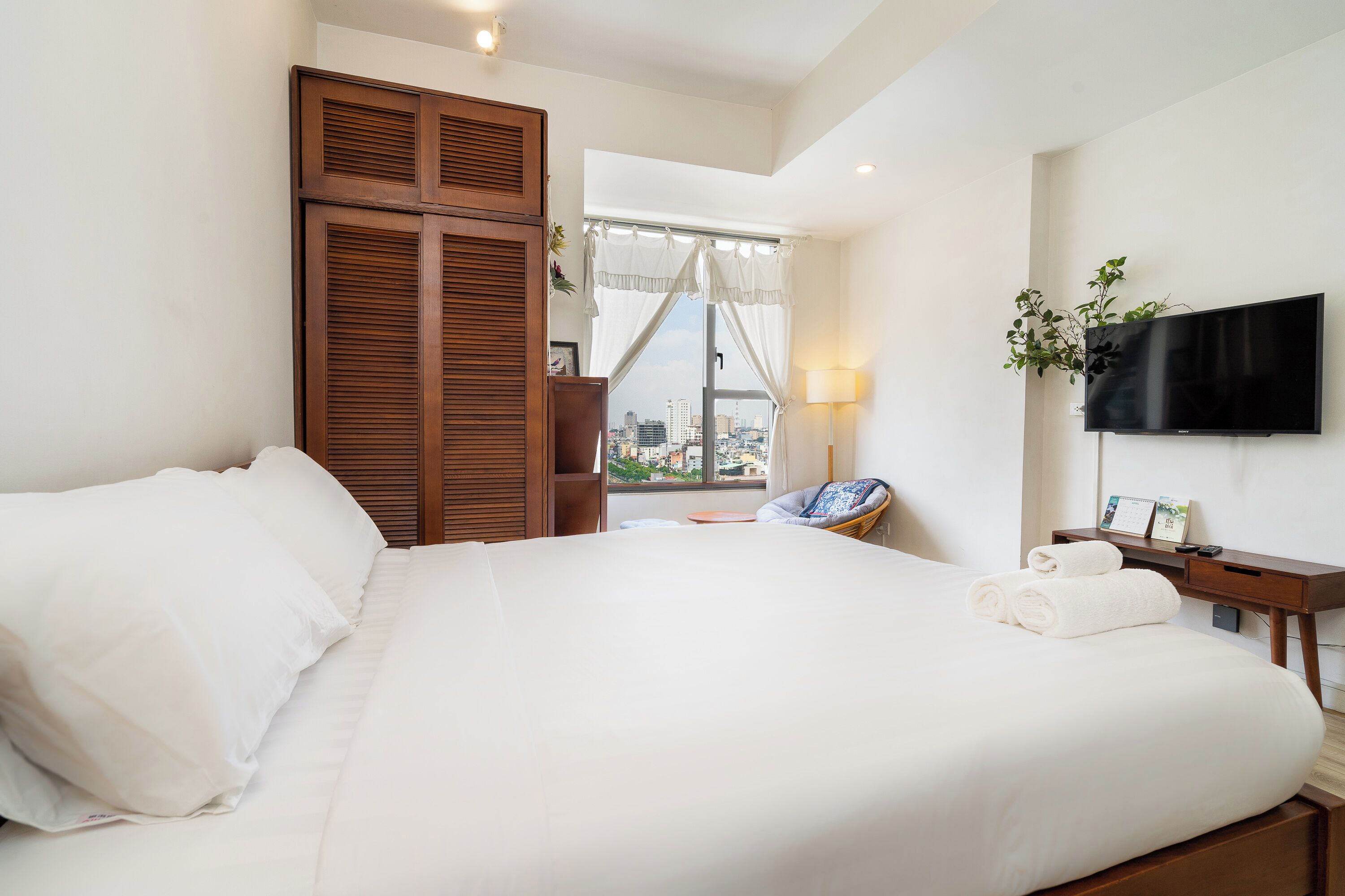 A neatly made bed with white linens, a wooden wardrobe, a bedside table with a lamp, and a TV mounted on the wall.