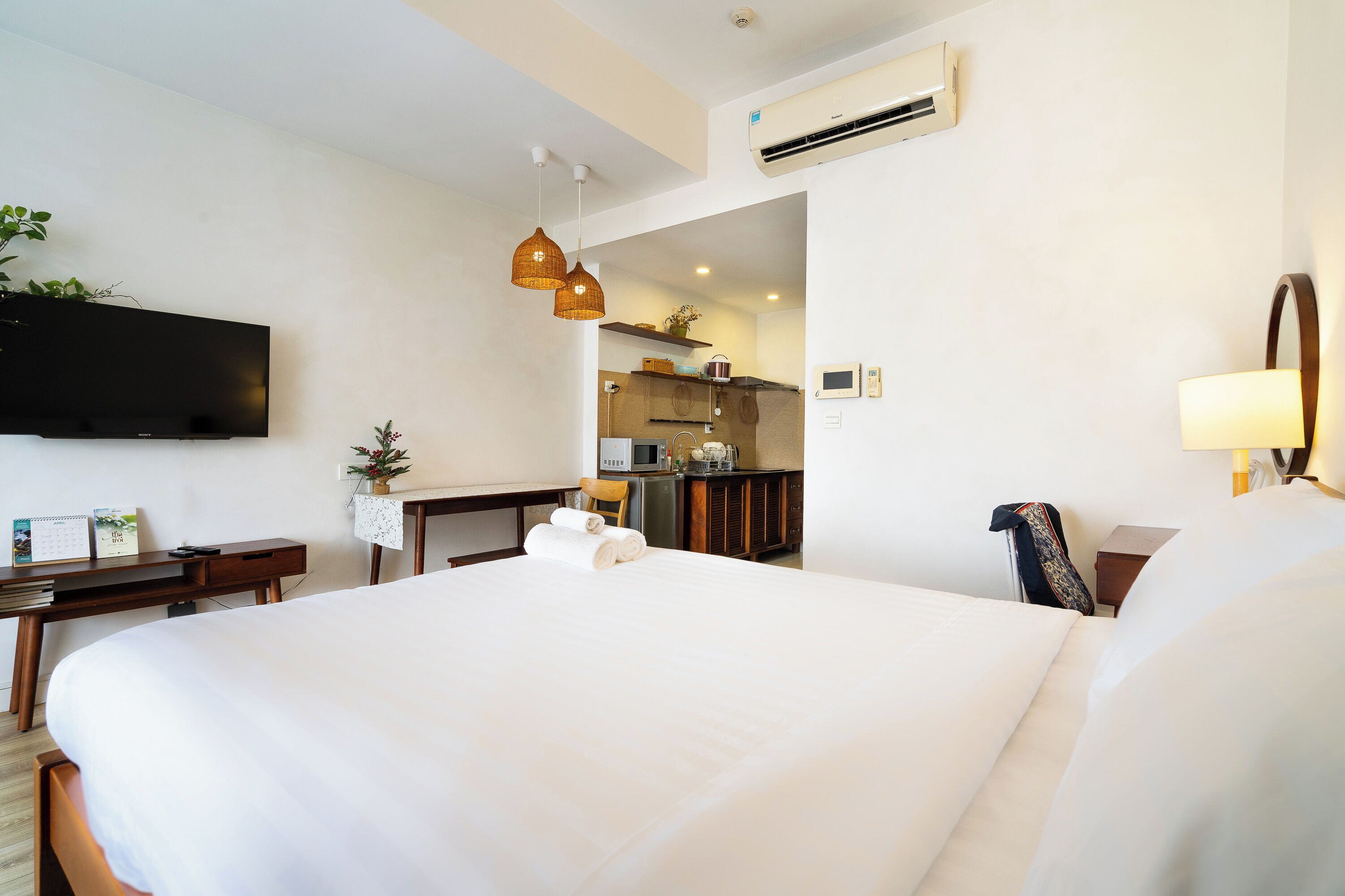 A hotel room with a large bed, a desk with a chair, a flat-screen TV mounted on the wall, and a kitchenette in the background.