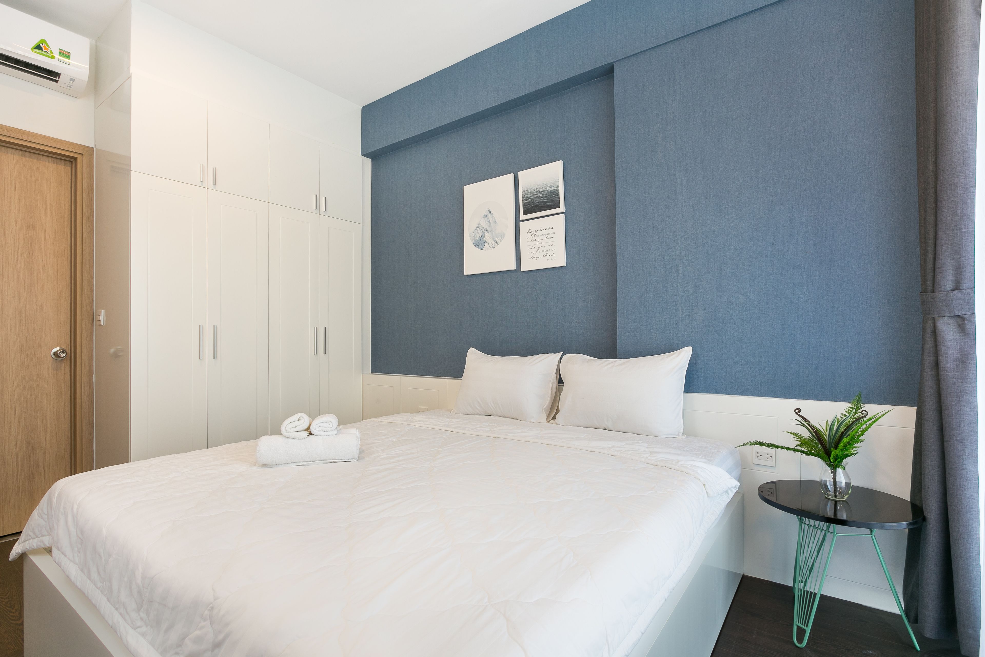 Una cama bien hecha con ropa blanca, una mesita de noche con una planta y una obra de arte colgada en la pared.