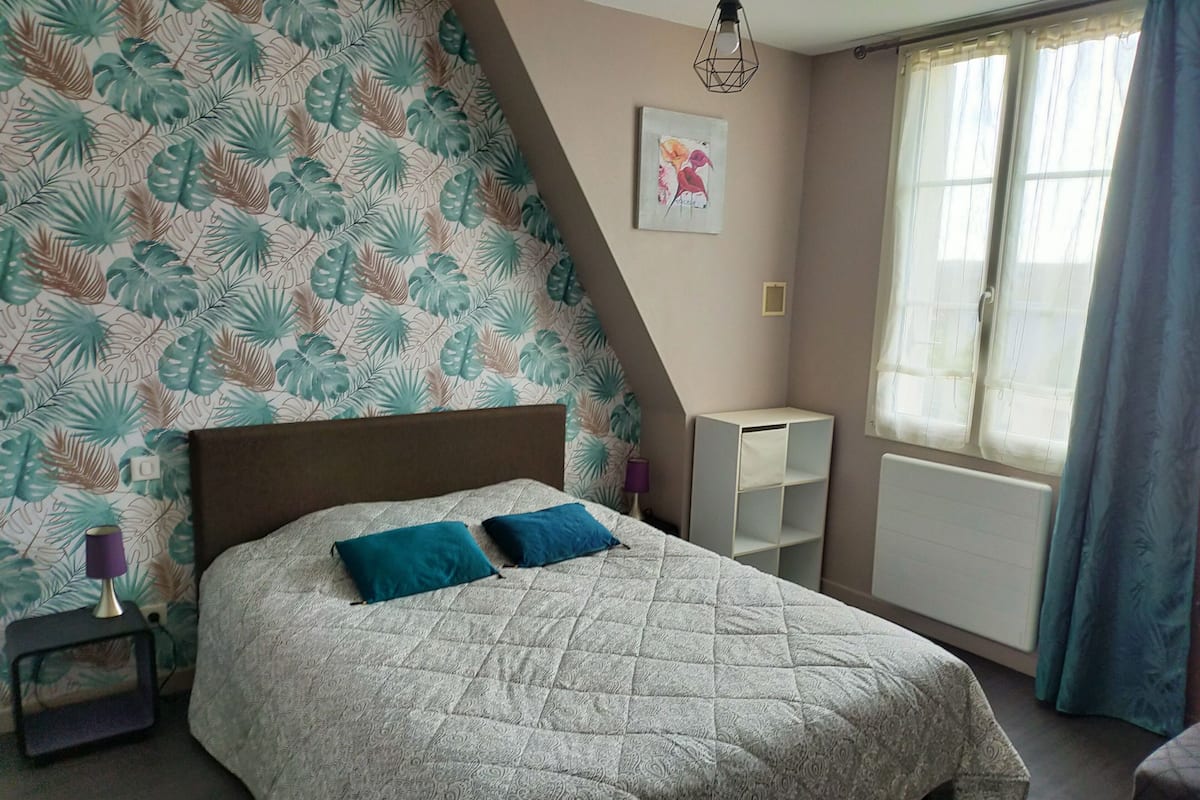 A bedroom with a bed, a nightstand, a wall-mounted shelf, and a window with curtains.
