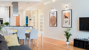 Ein modernes Wohnzimmer mit einer Couch, einem Esstisch und Stühlen. Im Hintergrund ist eine Küche mit Schränken und einem Herd zu sehen. An der Wand hängen zwei gerahmte Bilder, und ein Fernseher ist an der Wand befestigt.