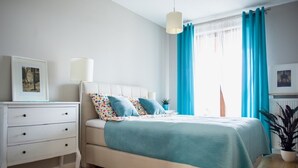 Ein Schlafzimmer mit Bett, Nachttisch, Lampe, Kommode und einem Fenster mit Vorhängen.