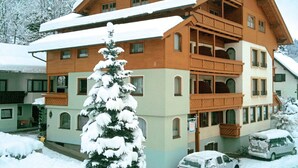 A multi-story wooden building with balconies, surrounded by snow-covered trees and parked vehicles. - Familienhotel Steindl (Millstatt)
