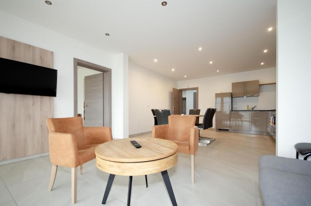 Een moderne woonkamer met een ronde houten tafel, twee leren fauteuils en een grijze bank. Een flatscreen televisie is aan de muur bevestigd.