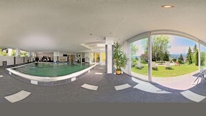 A modern indoor swimming pool with a view of a landscaped garden and a poolside area with lounge chairs. - Familienhotel Steindl (Millstatt)