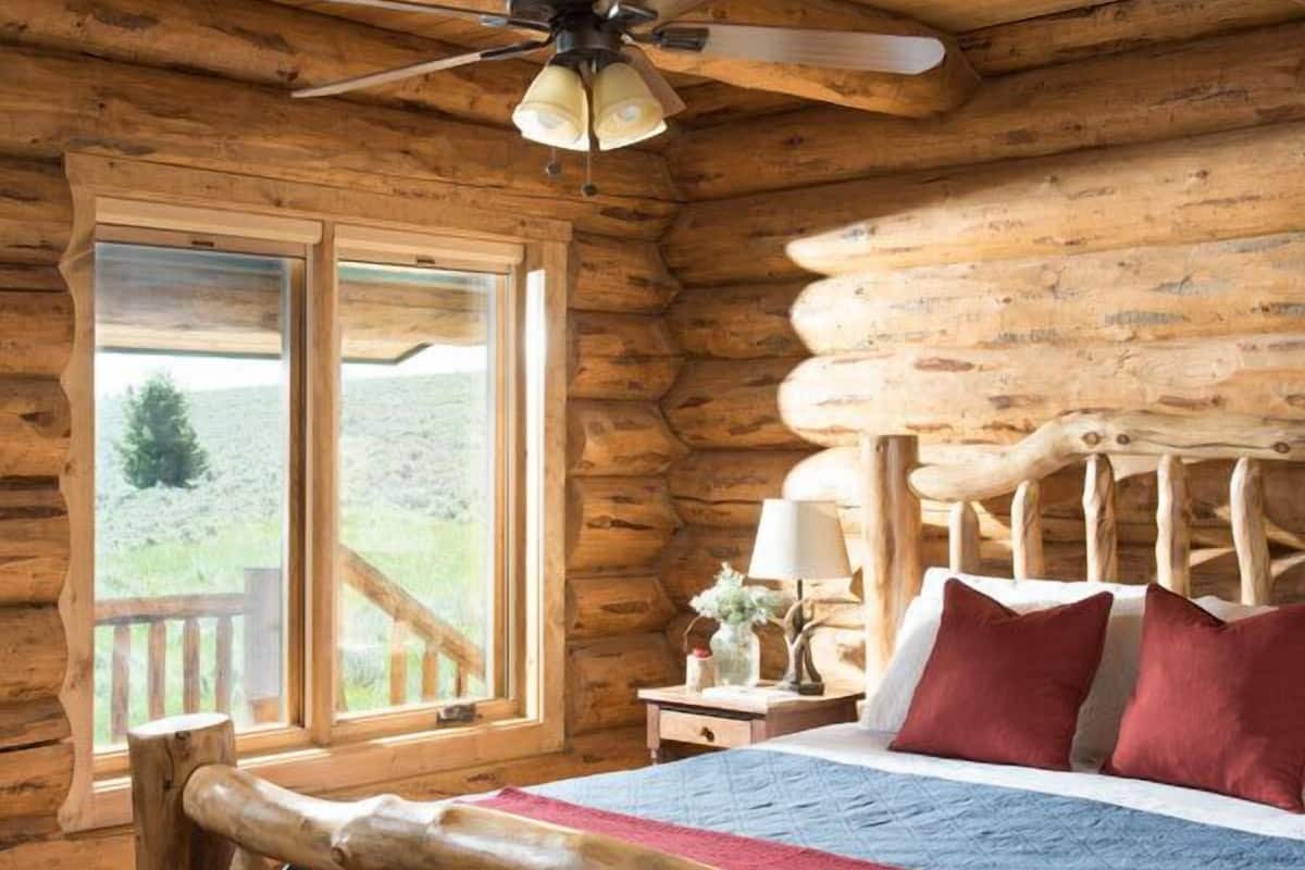 Une chambre située dans une cabane en rondins, avec un lit en bois, un ventilateur de plafond et une grande fenêtre donnant sur le paysage extérieur.