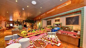 Un buffet proposant une variété de fruits, de desserts et un coin café, installé dans une pièce dont le plafond est en bois et les murs recouverts de carreaux verts.