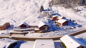 Un complejo turístico en la montaña cubierto de nieve, con varios edificios, incluido un hotel y un telesilla, rodeado de árboles nevados y una carretera.
