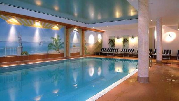 Une piscine intérieure ornée d’une fresque représentant un paysage tropical, avec des chaises longues et un plafond doté d’un éclairage encastré.