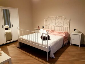 Een slaapkamer met een bed met wit metalen frame, een nachtkastje met een lamp, een kledingkast en een klein nachtkastje met een decoratief object.