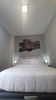 Una cama grande con colchón blanco y cabecera oscura, flanqueada por dos mesitas de noche con lámparas, en una habitación con una obra de arte grande colgada en la pared sobre la cama.