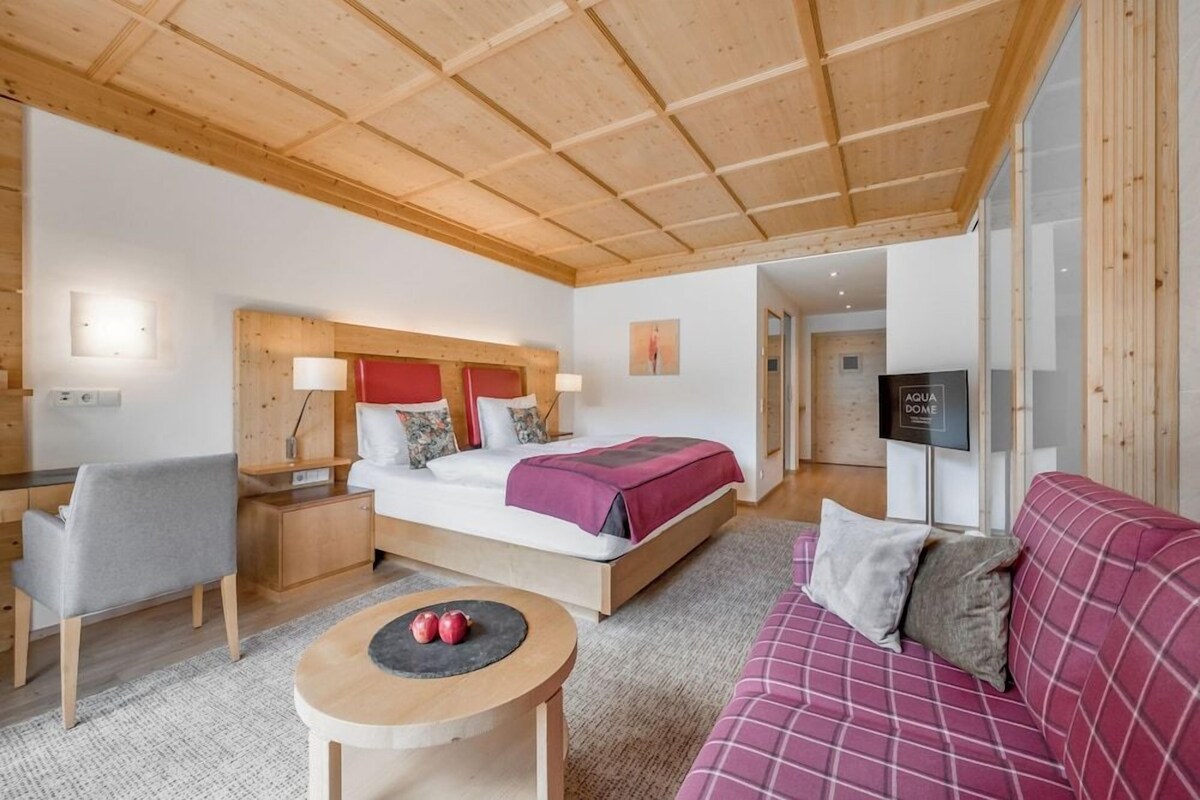 A modern hotel room with a wooden ceiling, a bed with a red headboard, a sofa, a desk, and a television.