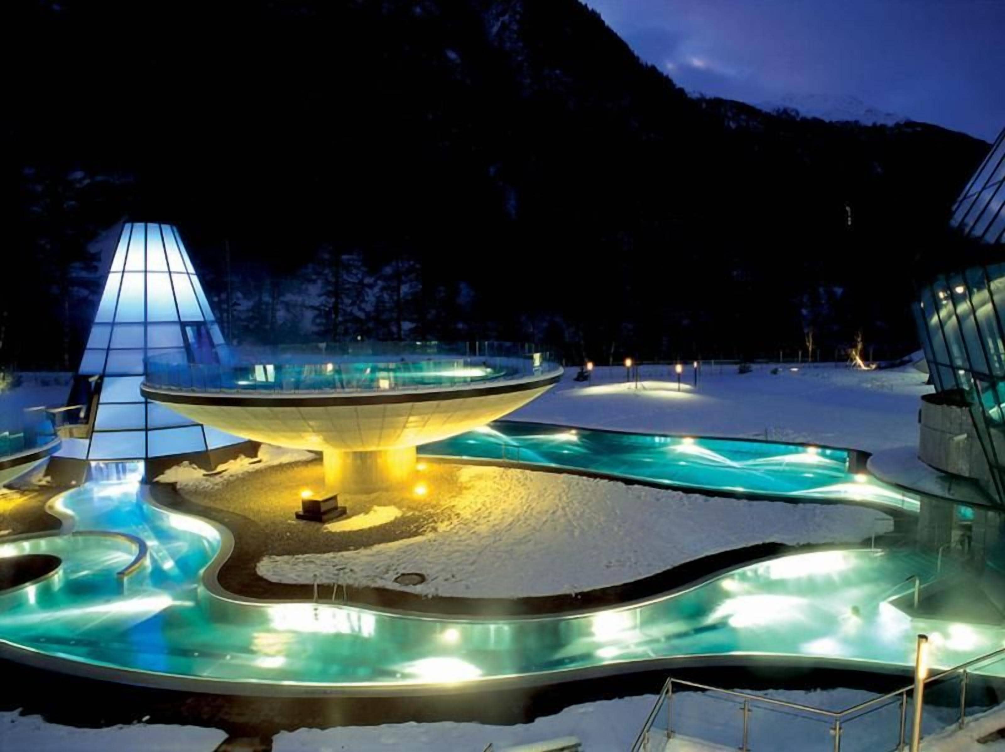Ein verschneiter Außenbereich mit einem zentral gelegenen, beheizten Pool, einer Glasstruktur und einer Bergkulisse.