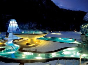A snow-covered outdoor area with a central heated pool, a glass structure, and a mountainous backdrop. - Aqua Dome Therme (Laengenfeld)