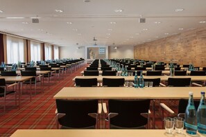 Un salón de conferencias con filas de mesas y sillas, botellas de agua y una pantalla para presentaciones.