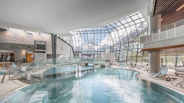 A large indoor swimming pool with a glass ceiling, surrounded by lounge areas and a stone wall. - Aqua Dome Therme (Laengenfeld)