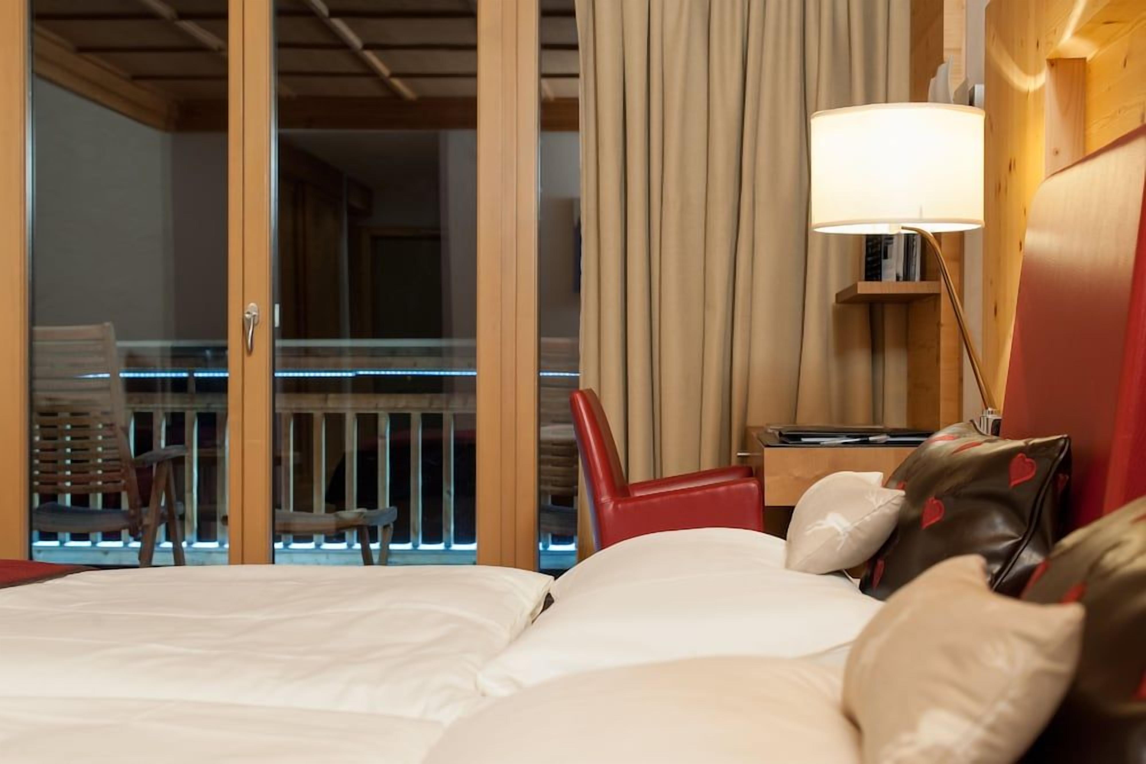 Ein Hotelzimmer mit einem Bett, einem roten Sessel, einer Lampe und Blick auf einen Balkon mit Lichtern.