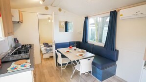 A compact kitchen with a gas stove, a dining table with blue cushions, and a window with curtains. - Happy Camp mobile homes in Füred Camping (Balatonfured)