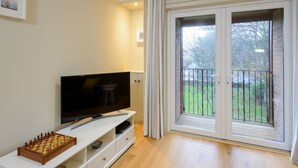 Un saló amb una televisió de pantalla plana sobre un moble blanc, un tauler d'escacs i una porta corredissa de vidre que dona a un balcó amb vistes a un jardí.