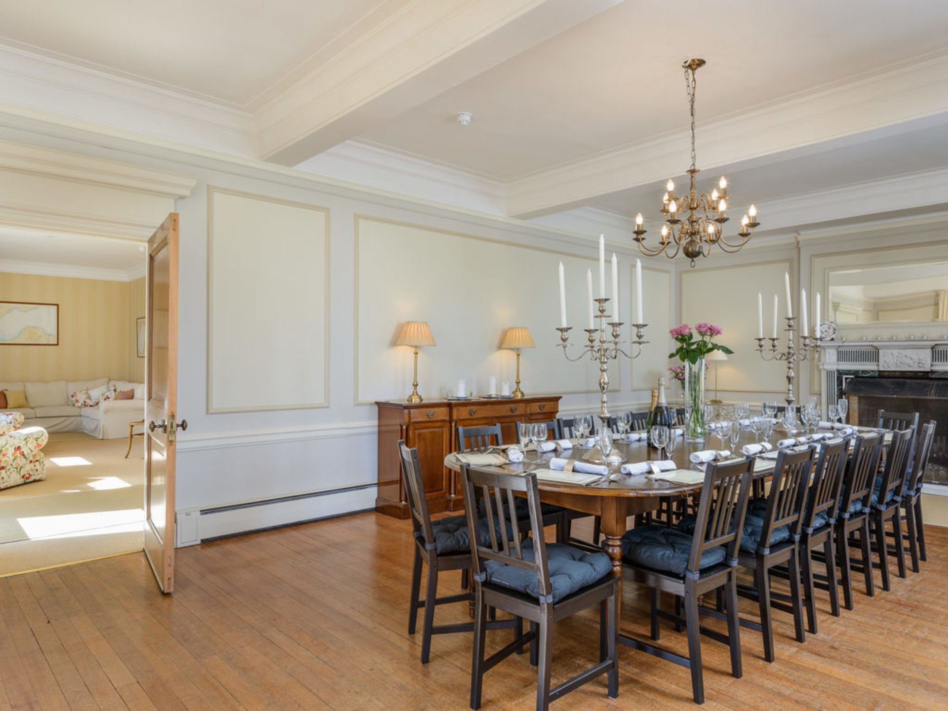 Uma sala de jantar formal com uma grande mesa de madeira posta para uma refeição, ladeada por cadeiras altas com assentos acolchoados. Um candelabro pendurado no teto, e uma lareira visível ao fundo.