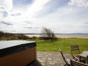 A patio with a hot tub, wooden chairs, and a view of a beach and distant mountains. - Green Craig (Aberlady)