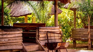 A rustic outdoor seating area with wooden benches and a metal chair, surrounded by lush greenery and thatched-roof structures. - Dream Valley Park & Lodge (Lusaka)